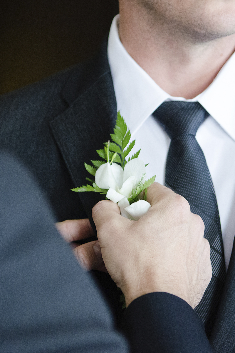 Detalle de mano de mujer colocando una flor blanca en el bolsillo del traje de novio de El Dique Moda Hombre